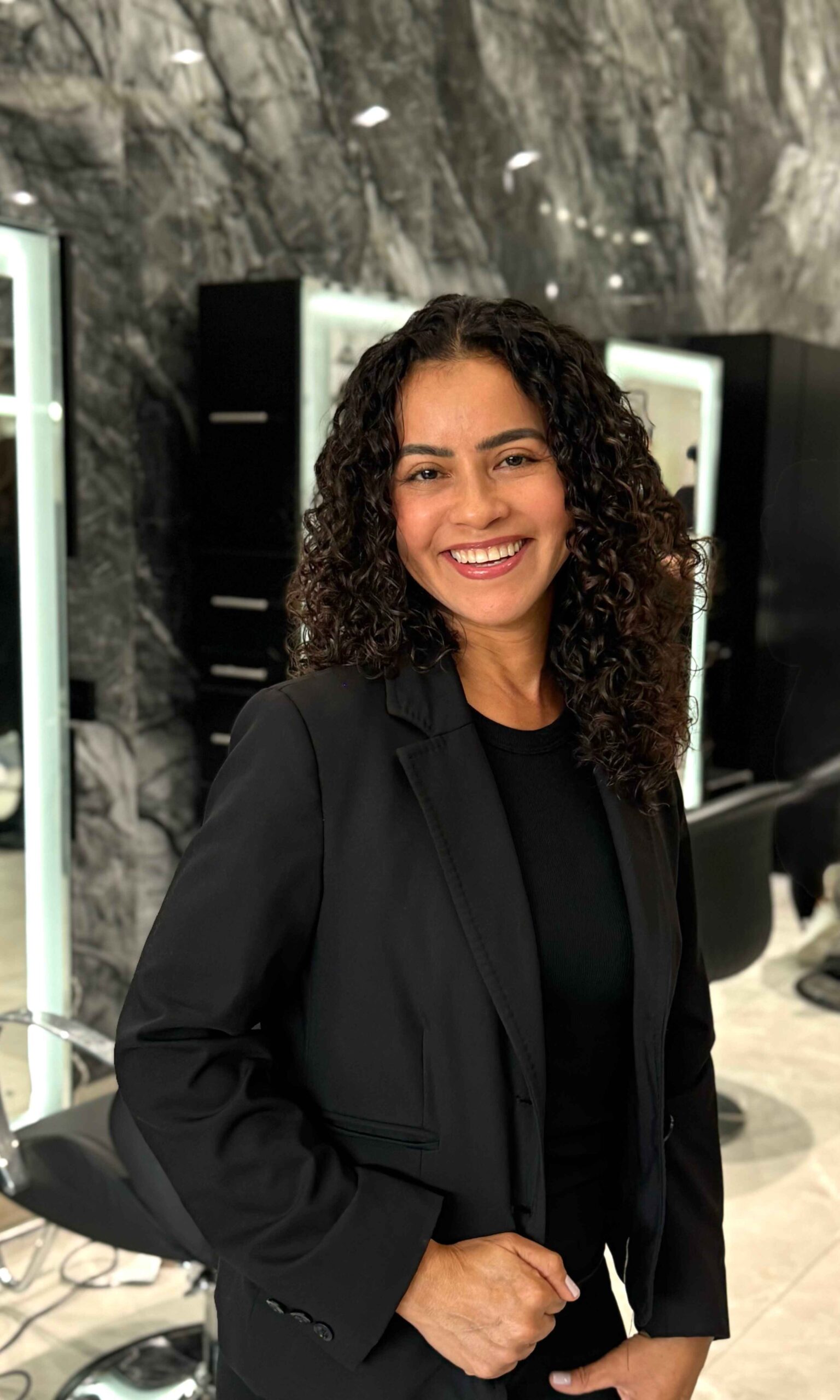 Vanesa our hairstylist she is smiling , she has curly hair and wearing a jacket with pants